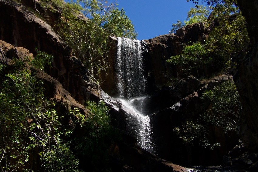 Waterfall cascading over rugged rock cliffs at Litchfield National Park in the Northern Territory, Australia. Surrounded by lush greenery, the waterfall drops into a shaded pool below, creating a serene and natural setting.