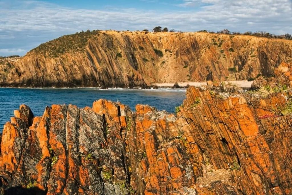 Rugged orange and grey rocks overlooking the secluded Little King George Beach on Kangaroo Island, with cliffs and a calm ocean in the background under a partly cloudy sky.