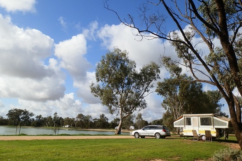 A caravan parked beside a car at Mallee Bush Retreat, with a grassy foreground and a view of Lake Lascelles. The sky is bright with fluffy clouds, giving a sense of a relaxing day at the campsite, one of the things to do in Hopetoun Victoria.