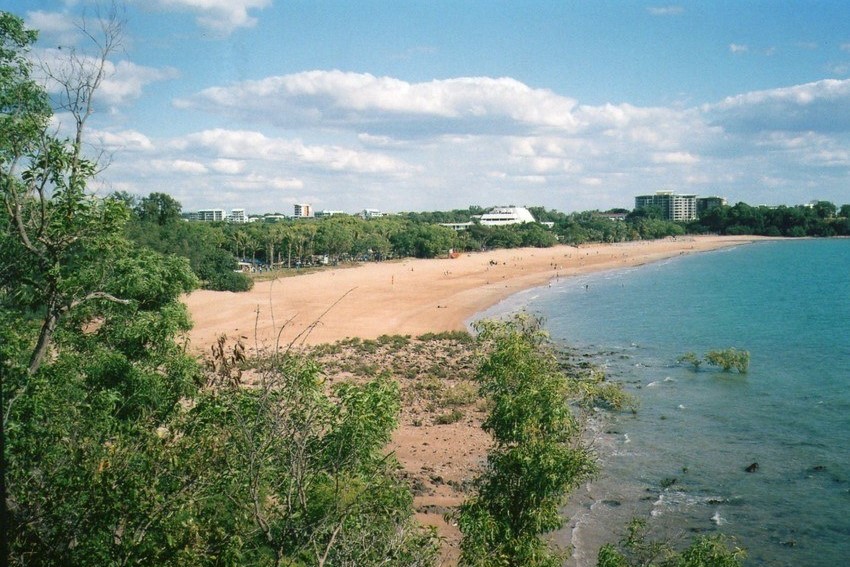 Mindil Beach in Darwin, Northern Territory, during the day. The sandy beach stretches along the shoreline with calm blue waters, while green trees frame the scene, and a few scattered buildings are visible in the background. The sky is mostly clear with some fluffy clouds, creating a peaceful beach atmosphere.
