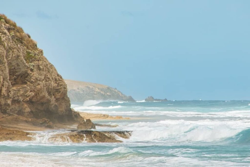 A scenic view of Mouth Flat Beach on Kangaroo Island, South Australia. The image shows a rocky cliff face on the left side of the frame, with the ocean in the background. The waves are crashing against the shore, creating a dramatic scene. The sky is a light blue, and the water is a turquoise color.