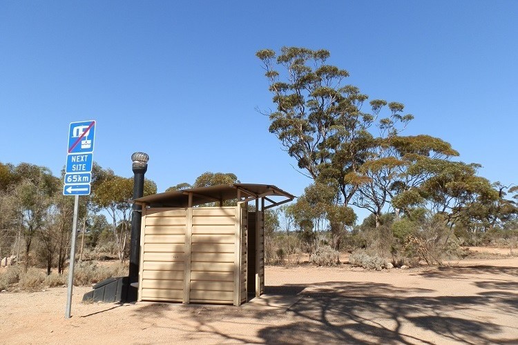 Basic rest stop facility along the Nullarbor Plain with a sign indicating the next rest area is 65 km away, surrounded by sparse trees and open landscape.