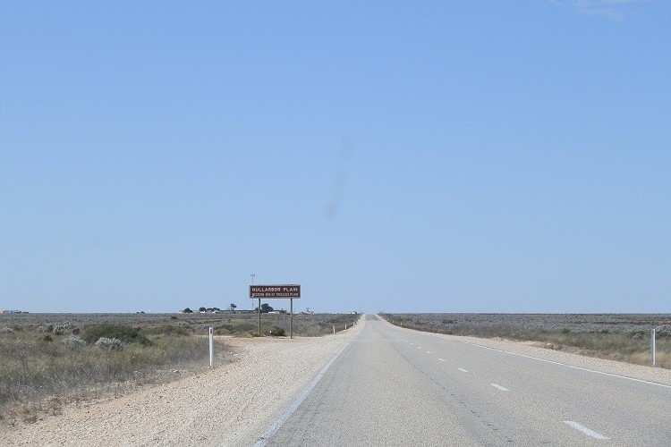 Long, straight road across the Nullarbor Plain with a sign marking its western edge, highlighting the vast, open landscape.