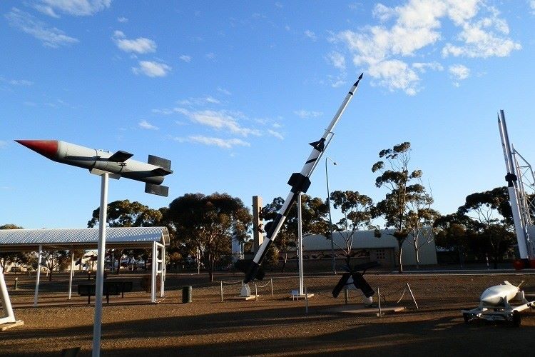 The Outdoor Missile Park in Woomera, on the way to Coober Pedy, South Australia, featuring various rockets and missiles on display. The exhibits are positioned on stands in an open area, with a clear blue sky and scattered trees in the background. The scene highlights the historical significance of the site as a hub for aerospace and military testing.