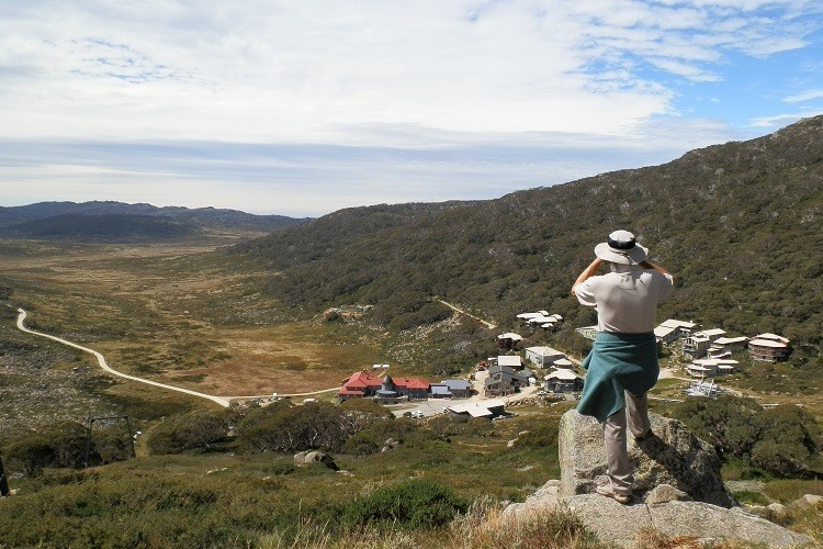 A person stands on a rock overlooking Charlotte Pass in New South Wales, capturing the view of the valley, winding path, and small buildings below, with mountains in the background.