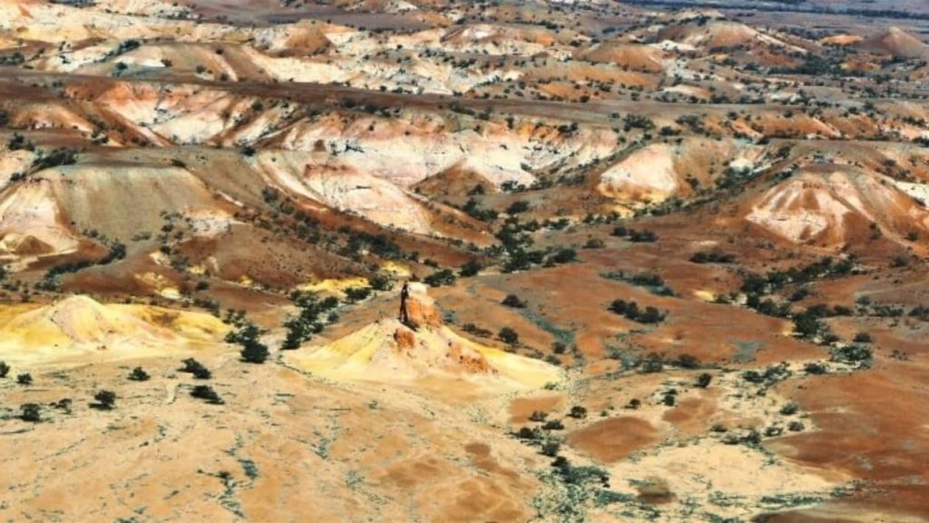 Aerial view of a distinct rock pillar standing amidst the colourful terrain of Anna Creek Painted Hills, in the outback of South Australia, surrounded by rolling mounds in shades of red, orange, and yellow.