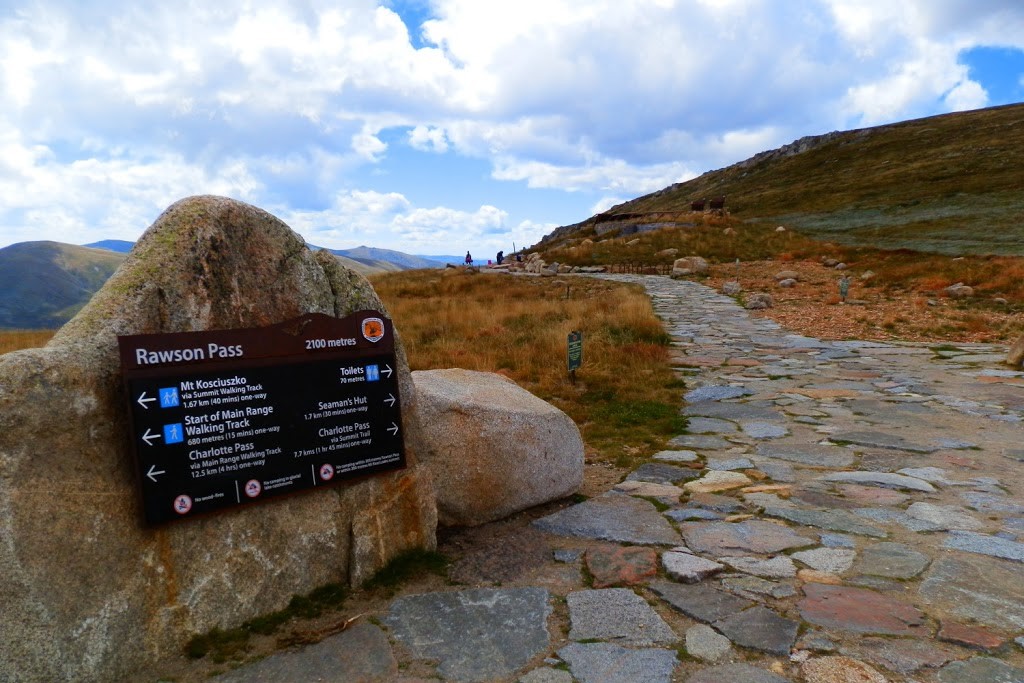Rawson Pass sign at 2100 meters, showing directions to highest toilet in australia, trails and landmarks, with a stone path and grassy hillside in the background.
