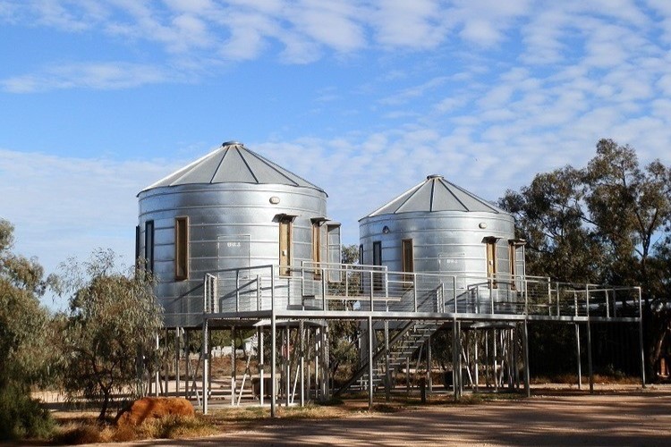 Two unique, cylindrical silo accommodations elevated on platforms at the Mallee Bush Retreat, surrounded by eucalyptus trees, under a clear, lightly clouded sky.