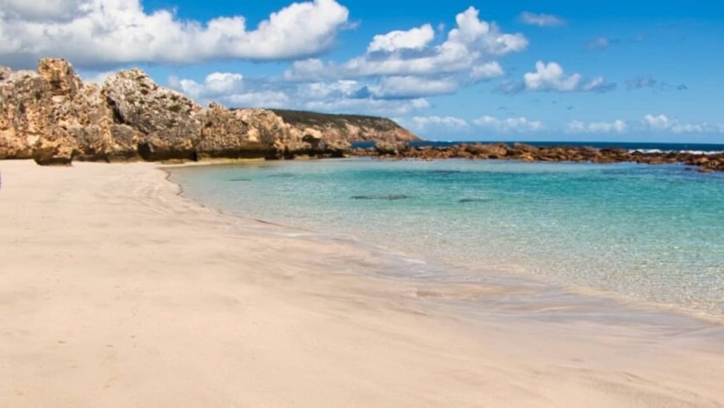 Crystal-clear waters and soft sandy shore of Stokes Beach, Kangaroo Island, with rugged rock formations and a blue sky filled with scattered clouds.
