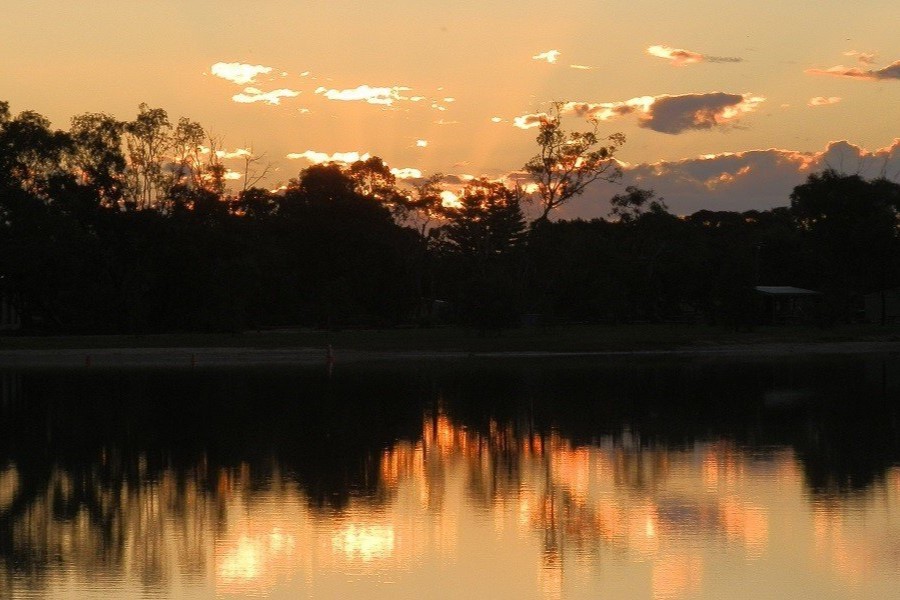 A serene sunset view over Lake Lascelles, with golden-orange light reflecting off the calm water and silhouettes of trees against the fading sky.