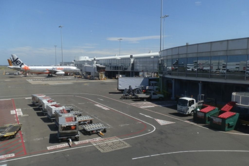 A Jetstar airplane is parked at the gate at Sydney Airport, surrounded by ground crew vehicles and luggage carts. The airport terminal building with large reflective windows can be seen in the background under a clear blue sky.