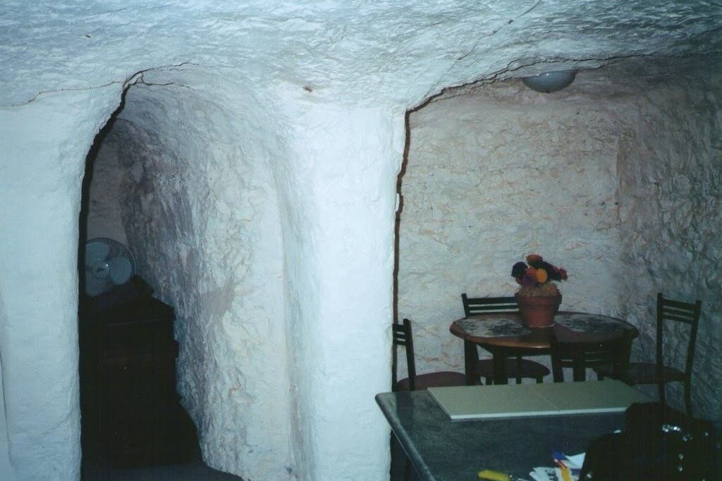 The interior of an underground accommodation in Coober Pedy, South Australia. The walls and ceiling are carved out of rock, with a rough, textured appearance typical of the region's dugout homes. A small dining area with a table and chairs is visible, emphasizing the cozy and unique living environment found in this opal mining town known for its subterranean lifestyle.
