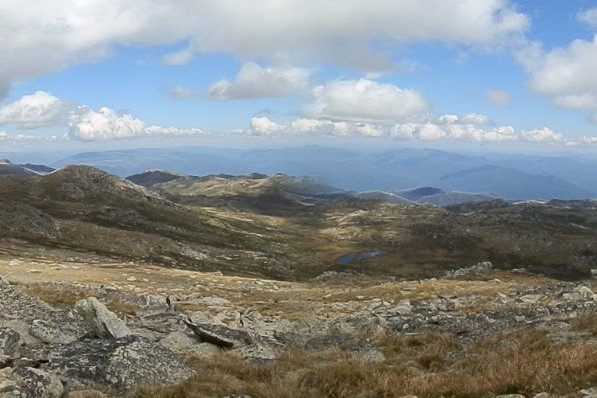 A panoramic view from Mt Kosciuszko in the Snowy Mountains, New South Wales, featuring rocky terrain, grassy slopes, and distant hills under a partly cloudy sky.