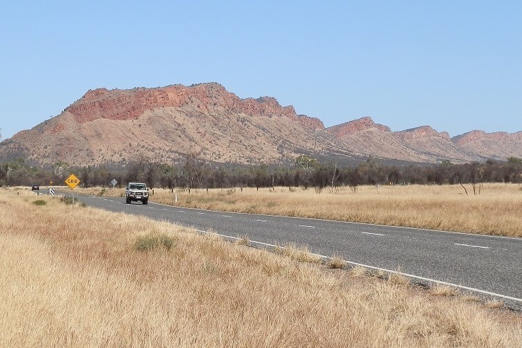 The Stuart Highway with the West MacDonnell Ranges in the background, in the Northern Territory of Australia. A vehicle is driving on the road, surrounded by dry grassland, with the rugged, reddish rock formations of the ranges dominating the background. The clear blue sky emphasizes the arid landscape and the natural beauty of the region.