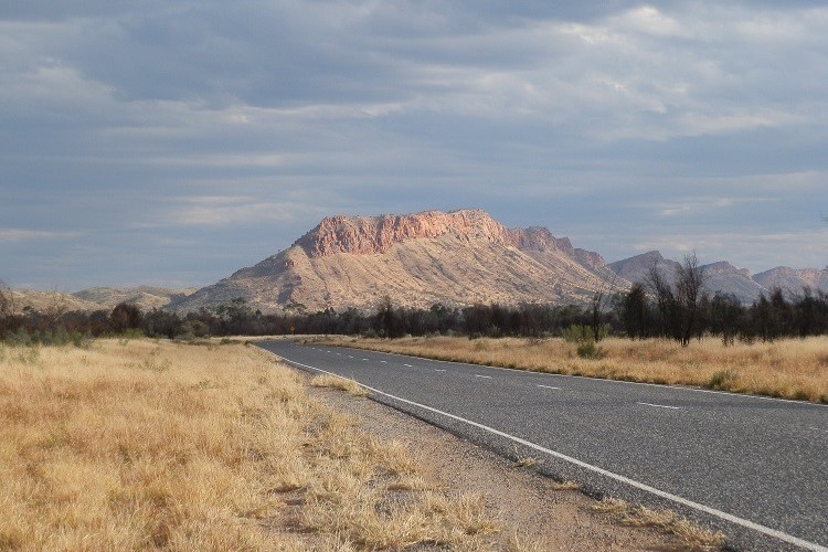 The image depicts the Stuart Highway, the main way to get from Adelaide to Darwin, with the West MacDonnell Ranges in the background, near Alice Springs in the Northern Territory, Australia. The rugged mountain range, characterized by its reddish rock formations, rises prominently in the distance under a cloudy sky. The roadside is lined with dry grass, highlighting the arid landscape typical of the region.