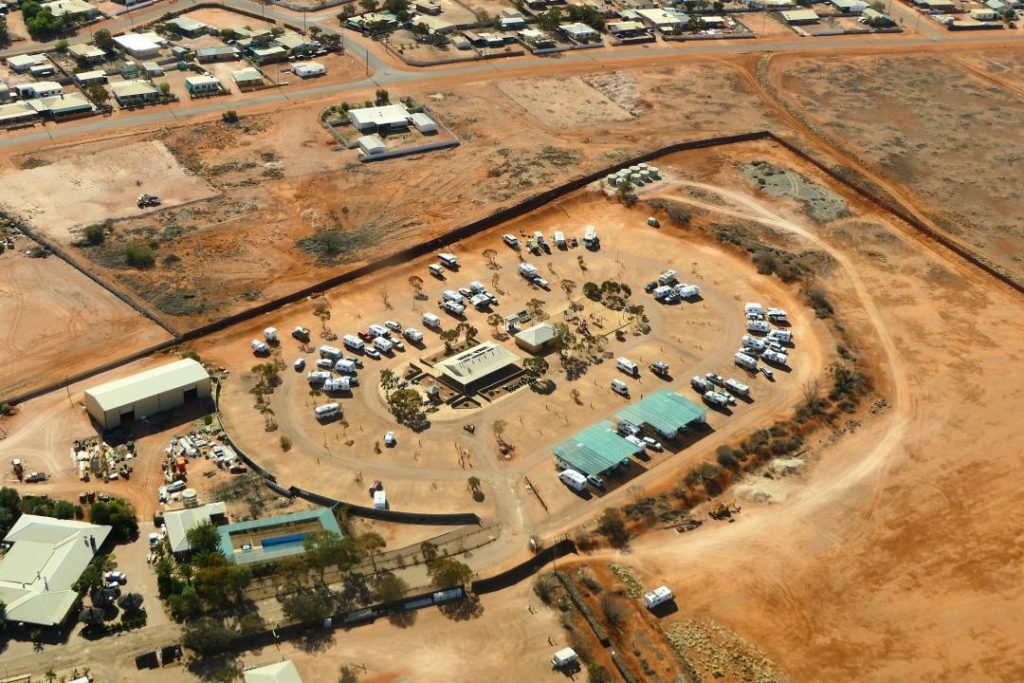 Caravan park in Coober Pedy, South Australia, surrounded by arid landscape. The park features numerous RVs and caravans parked in a circular arrangement around central facilities, with sparse vegetation scattered throughout. The surrounding area showcases the town's typical outback terrain, characterized by red earth and minimal greenery.
