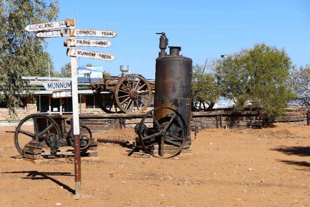 Vintage machinery and a signpost showing distances to various cities at William Creek, South Australia, highlighting the remote outback location under a clear blue sky.