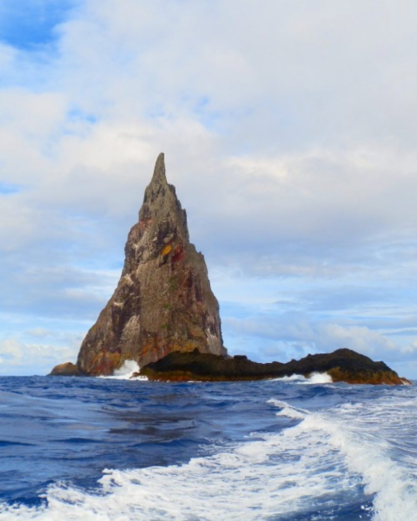 A tall, jagged, and isolated rock formation known as Balls Pyramid rises sharply from the ocean, surrounded by choppy waves.