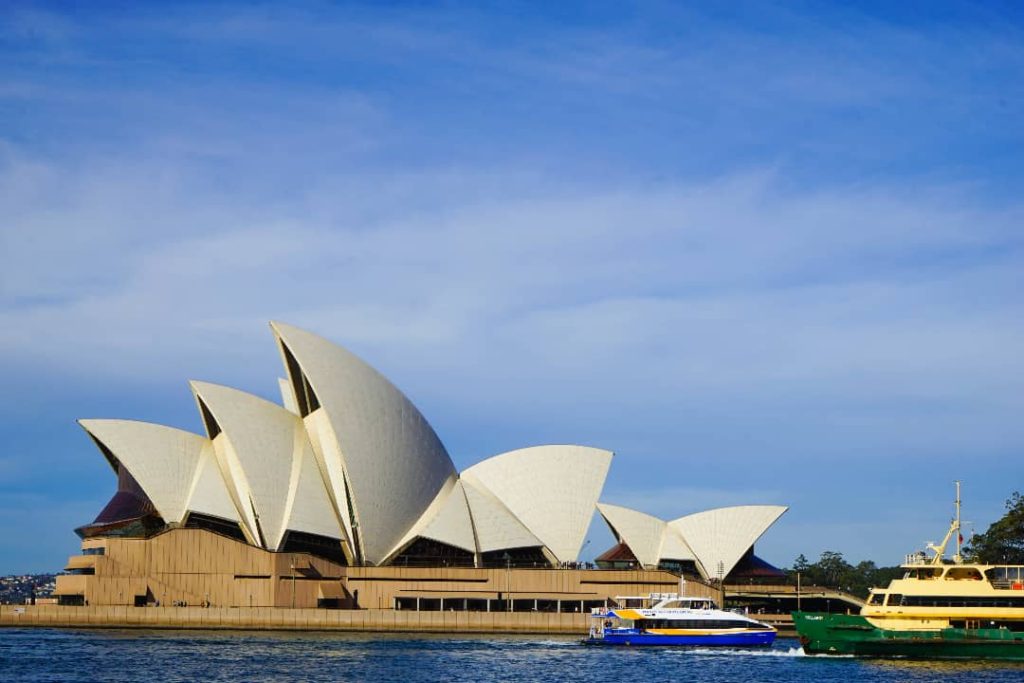 The Sydney Opera House stands proudly against a clear blue sky, its white sails gleaming in the sunlight. Boats, including a yellow ferry and a smaller vessel, pass in front of this iconic structure on the water.
