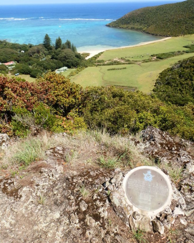 A memorial plaque at the Catalina plane crash site on Lord Howe Island, Australia, overlooks a scenic view of the island's lush landscape and the turquoise waters