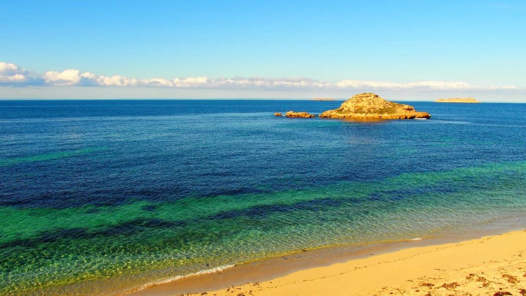A serene view of Chinaman's Hat Island, a rocky outcrop rising from the calm blue waters off the coast of Innes National Park, Yorke Peninsula, South Australia. The sandy beach in the foreground contrasts beautifully with the deep blues and greens of the ocean, while the distant horizon is lined with soft clouds.