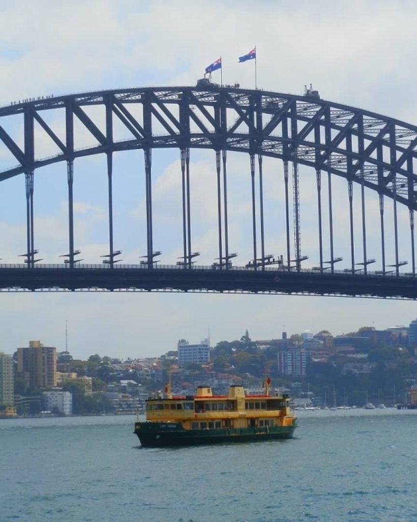 A ferry sails across Sydney Harbour with the iconic Sydney Harbour Bridge towering in the background. Tiny figures of climbers can be seen walking along the top arch of the bridge, flanked by two Australian flags waving in the breeze.