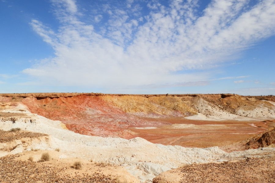 A vast, arid landscape at the Ochre Pits in Central Australia, featuring striking layers of ochre, red, yellow, and white soil. Wispy white clouds stretch across a clear blue sky, enhancing the natural beauty and highlighting the colorful, layered earth.