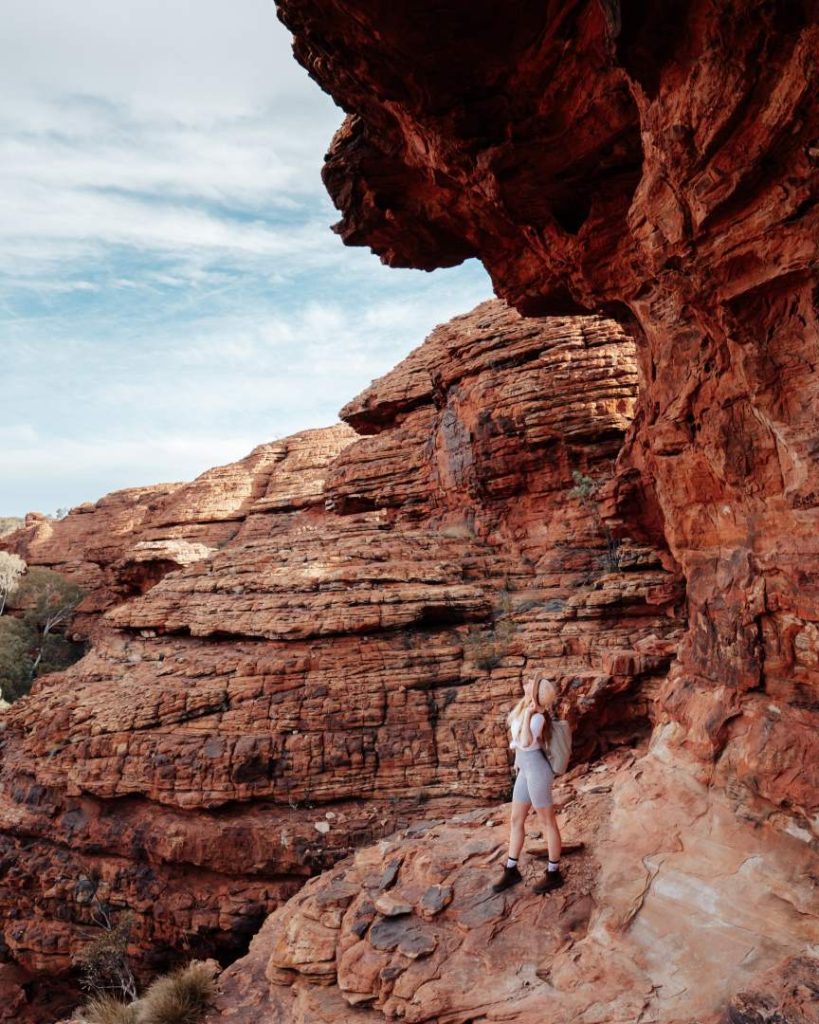 A woman hiking along the rugged rock formations of Kings Canyon in the Northern Territory, Australia. She is standing on a rocky ledge, surrounded by layered red cliffs that rise dramatically against the sky. The scene highlights the adventurous nature of the canyon's landscape and the striking geological features found in this iconic outback destination.