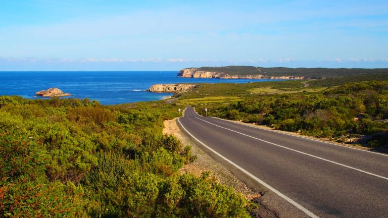 A scenic road stretches through the green coastal landscape of Innes National Park on the Yorke Peninsula, South Australia. The road leads towards the sparkling blue ocean, framed by rugged cliffs and small rocky islands.