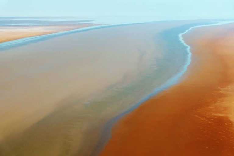 Aerial view of Lake Eyre in the outback of South Australia, showcasing colourful, smooth, and reflective salt flats with soft hues of orange, brown, and blue blending together.