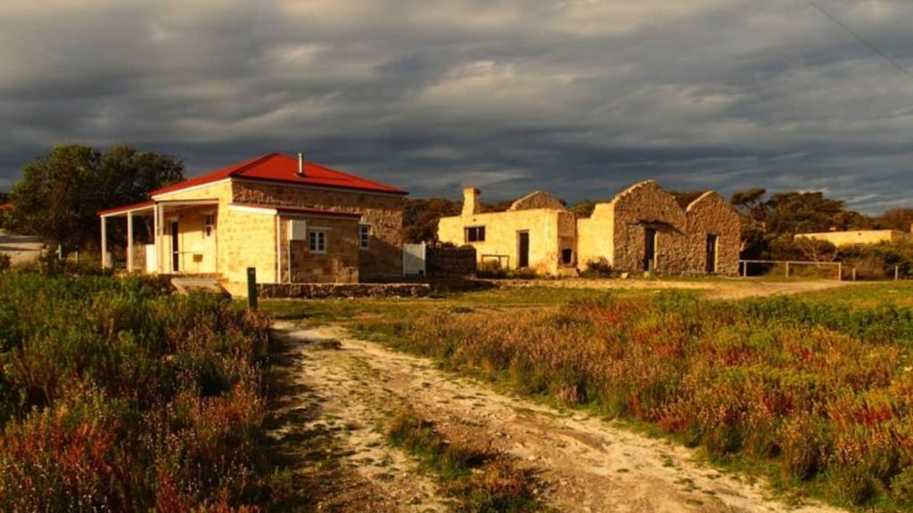 A heritage stone building with a bright red roof sits alongside the ruins of an older structure, both bathed in the golden light of the late evening in Inneston, Innes National Park, South Australia. The sky above is cloudy and dark, contrasting with the illuminated foreground of wild grasses.