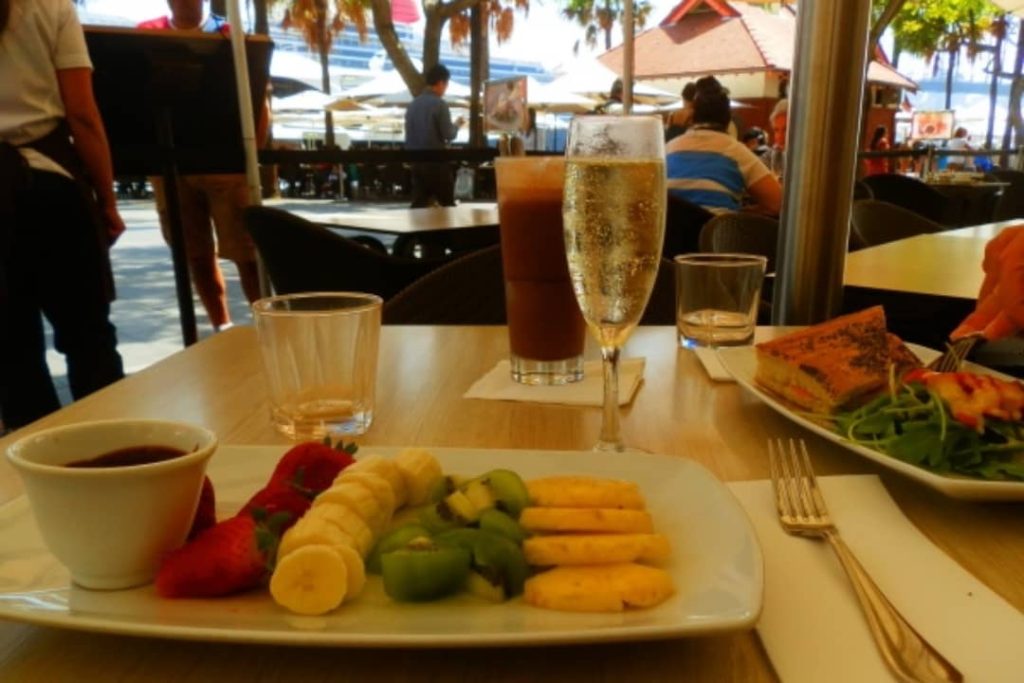 A café table at East Circular Quay, Sydney Harbour, features a plate of fresh fruit, a glass of champagne, and an iced coffee. In the background, people are enjoying the outdoor dining area with a view of Sydney Harbour.