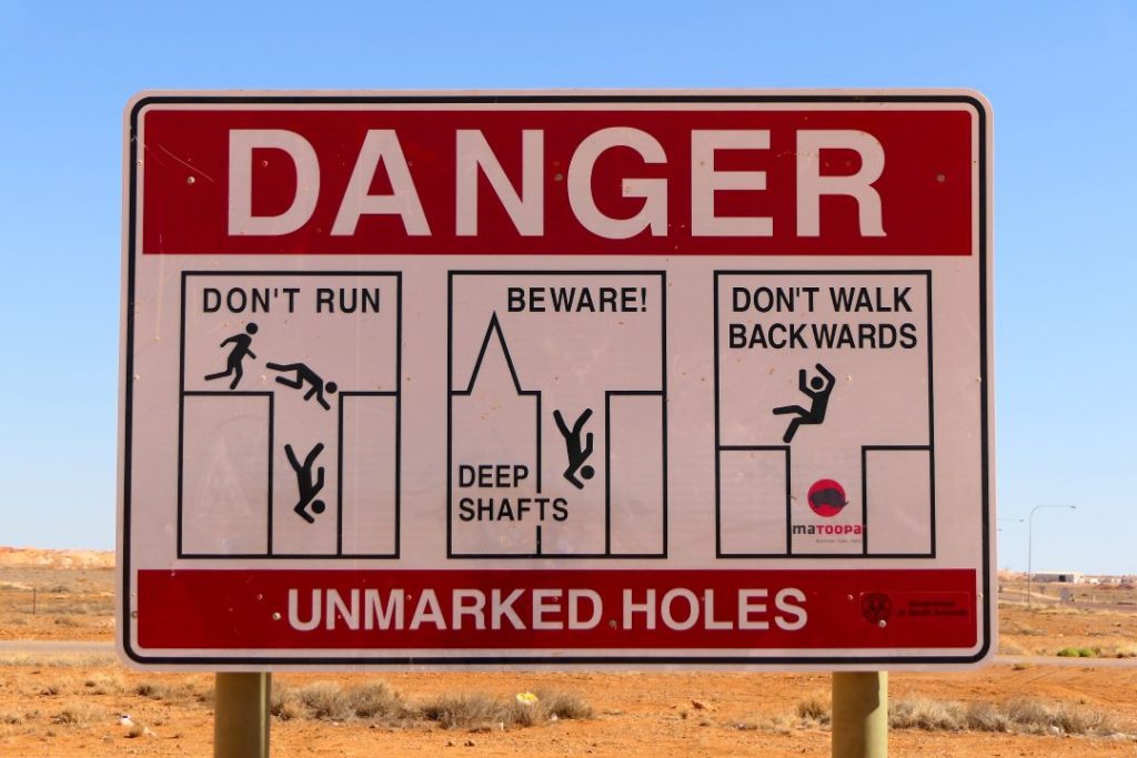 The image displays a warning sign in Coober Pedy, South Australia, cautioning about unmarked mining holes. The sign reads "DANGER" and features illustrations with the messages "DON'T RUN," "BEWARE! DEEP SHAFTS," and "DON'T WALK BACKWARDS," depicting stick figures falling into deep shafts. The background shows the arid landscape typical of the region, emphasizing the risks associated with the area's numerous open mining pits.