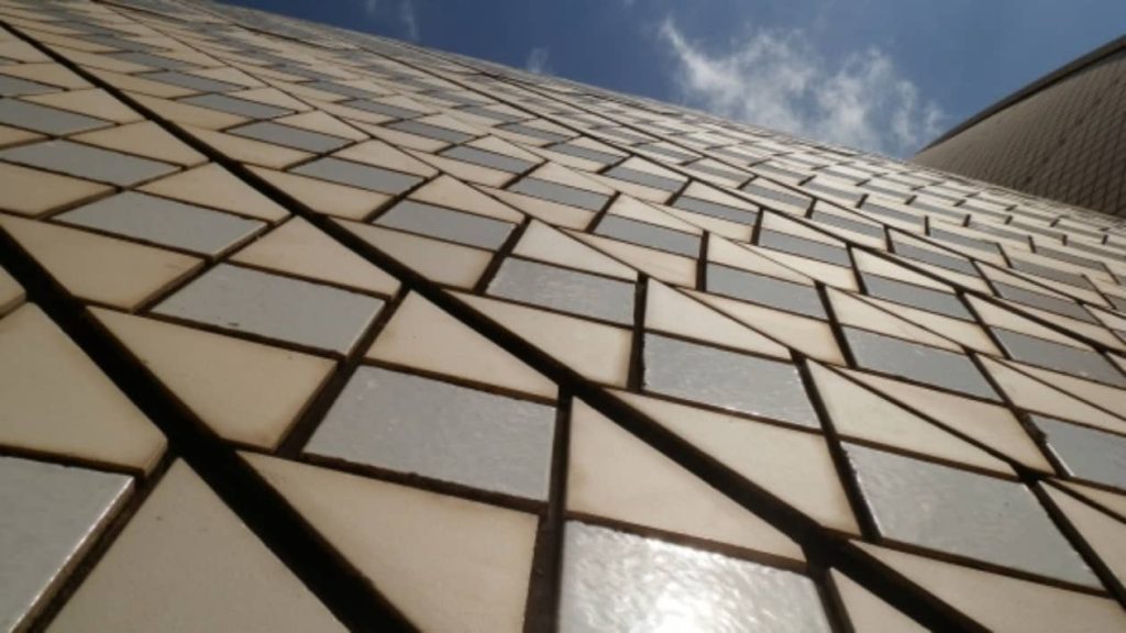 A close-up view of the Sydney Opera House's iconic white and cream tiles, showcasing the geometric pattern of interlocking triangles against the blue sky. The tiles' reflective surface gives them a glossy, textured appearance as they curve up the structure.