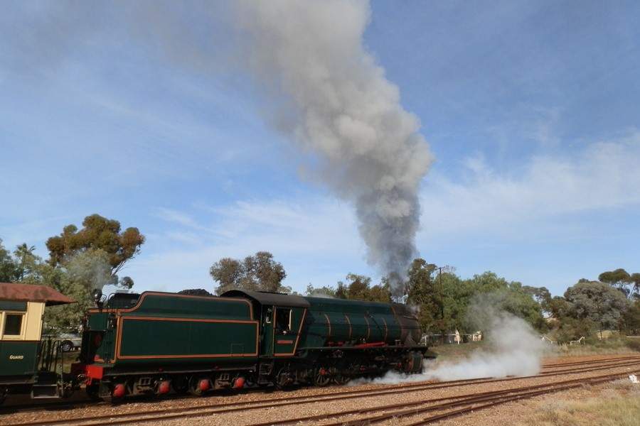A steam locomotive from the Pichi Richi Railway departing from Quorn Station in South Australia. The vintage train emits a thick plume of smoke as it travels along the railway tracks, with surrounding trees and a clear sky in the background. The scene captures the nostalgia and charm of historic rail travel.