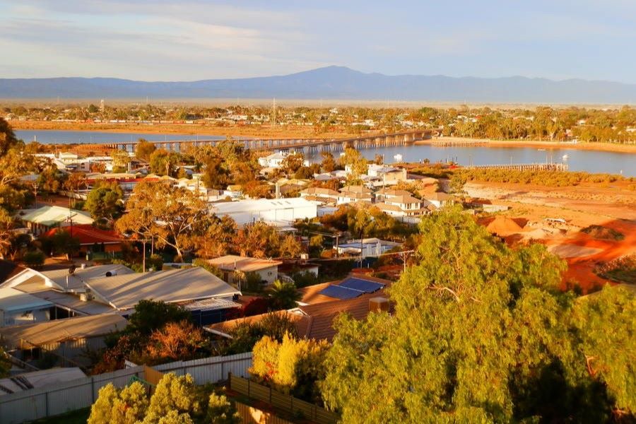 Aerial view of Port Augusta, South Australia, showing residential areas, greenery, and the Spencer Gulf in the background. The landscape includes a mix of trees, rooftops, and the distant mountain range under a softly lit sky, creating a warm, late afternoon atmosphere.