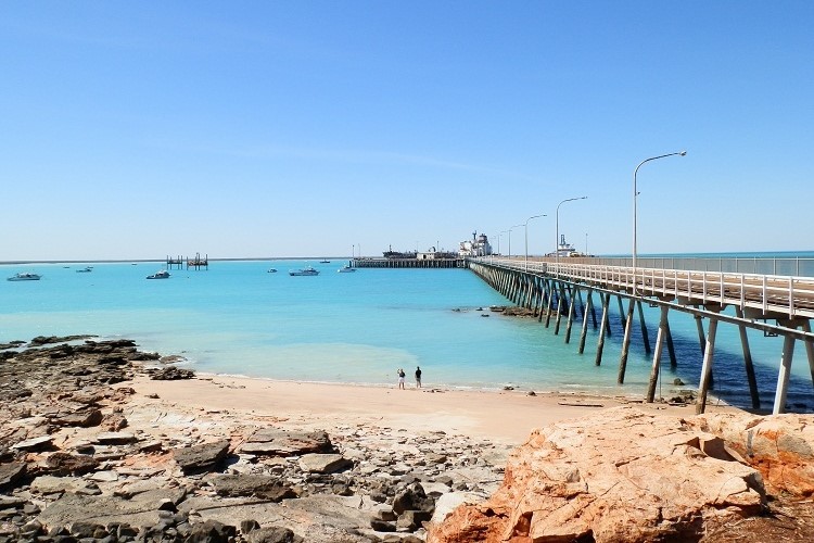 Port of Broome in Western Australia, featuring a long pier extending into the turquoise waters of the Indian Ocean. The beach in the foreground has light-colored sand and some rocky areas, while boats can be seen anchored in the distance. The clear blue sky and bright sunlight add to the scenic coastal atmosphere.