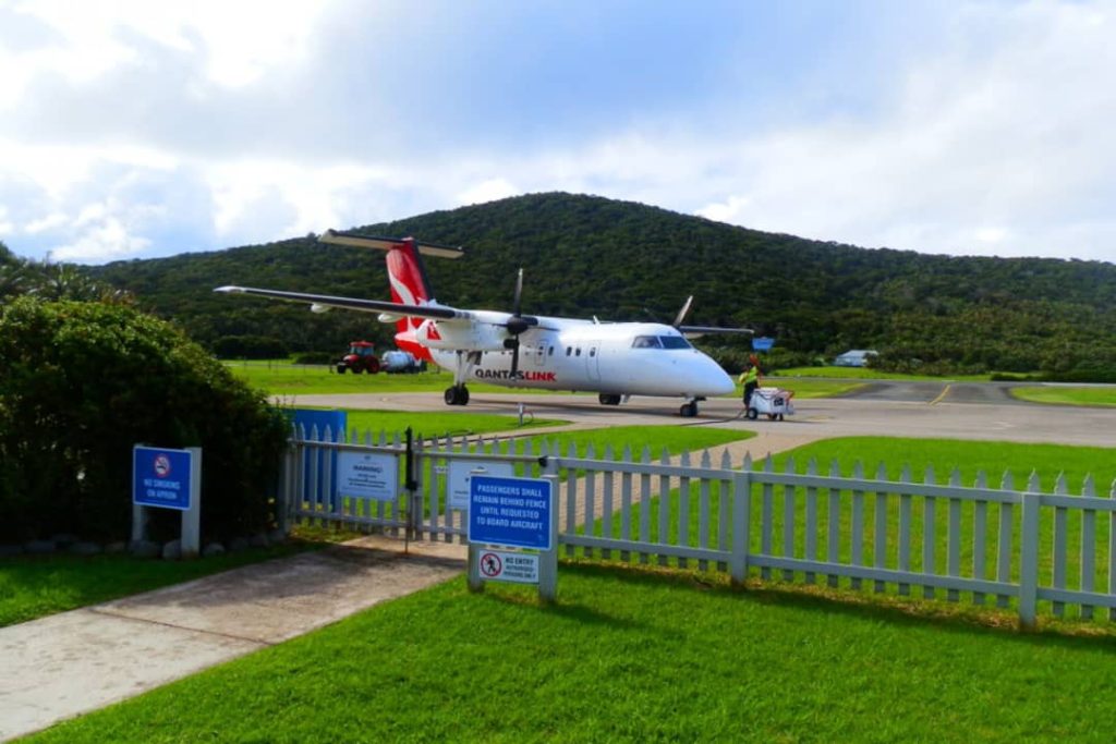 A QantasLink plane is parked on the airstrip at Lord Howe Island, surrounded by green hills and clear skies. The small airstrip allows passengers to fly directly to this remote and beautiful destination.