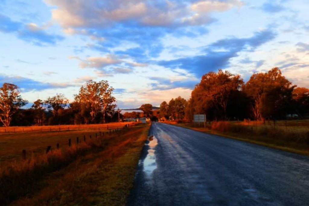 A scenic country road leading to Woodenbong, NSW, with trees and fields glowing under a beautiful sunset.