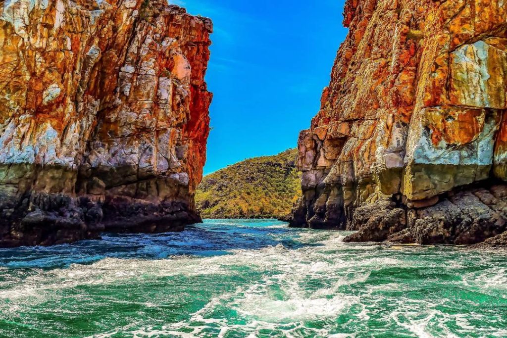 Dramatic view of the Horizontal Falls in the Kimberley region of Western Australia. Turquoise waters rush between two towering, rugged rock cliffs, creating a dynamic seascape under a clear, vibrant blue sky. The texture and color of the rocks, combined with the turbulent water, emphasize the natural beauty and power of this iconic geological formation.