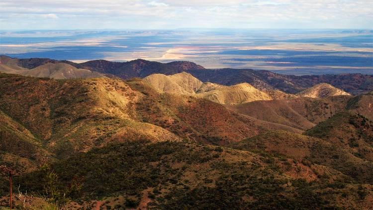 The image captures a summit view from Dutchmans Stern in the Flinders Ranges, South Australia. The landscape features rolling, rugged hills covered in sparse vegetation, with the distant plains stretching out towards the horizon. The scene is bathed in natural light, highlighting the varied textures and elevations of the terrain under a partly cloudy sky.