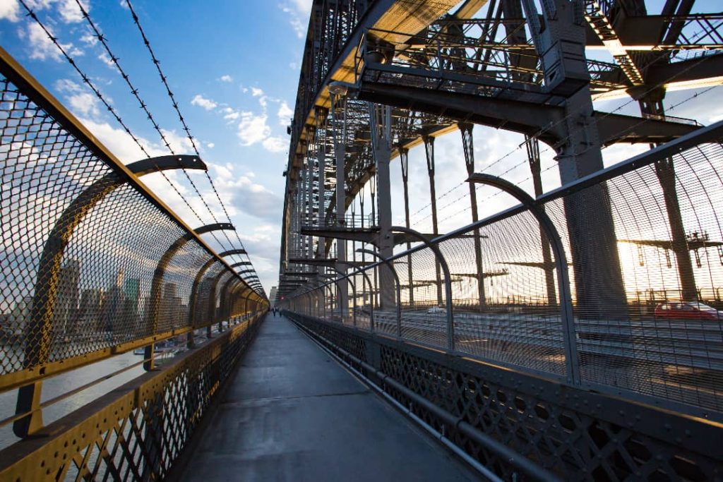 A pedestrian walkway along the Sydney Harbour Bridge, enclosed by metal fencing on both sides. The sun is setting in the background, casting a warm glow on the bridge's steel structure, with the Sydney skyline visible in the distance.