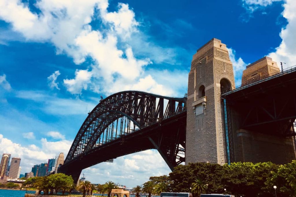 A close-up view of the Sydney Harbour Bridge on a sunny day with blue skies and scattered clouds. The iconic steel structure looms above, with the bridge's massive stone pillars prominently visible.
