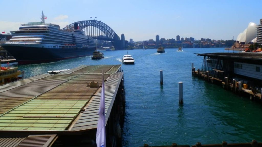 This image captures a view of Sydney Harbour from Circular Quay, featuring the Sydney Harbour Bridge in the background and a large cruise ship docked nearby. Several boats and ferries move across the water, with the Sydney Opera House visible on the right under a clear, sunny sky.