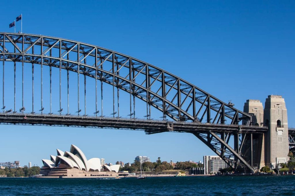 The Sydney Harbour Bridge stands tall against a clear blue sky, with the iconic Sydney Opera House visible in the background. The waters of the harbour shimmer under the bright sunlight, while the Australian flags wave at the top of the bridge.