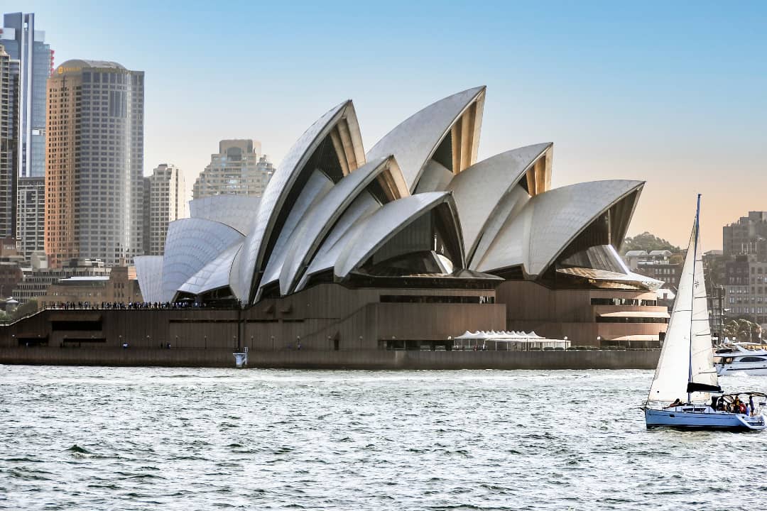 The Sydney Opera House stands gracefully against the backdrop of the city skyline during sunset. A sailboat drifts in the foreground on the calm waters, adding to the peaceful and iconic scene.