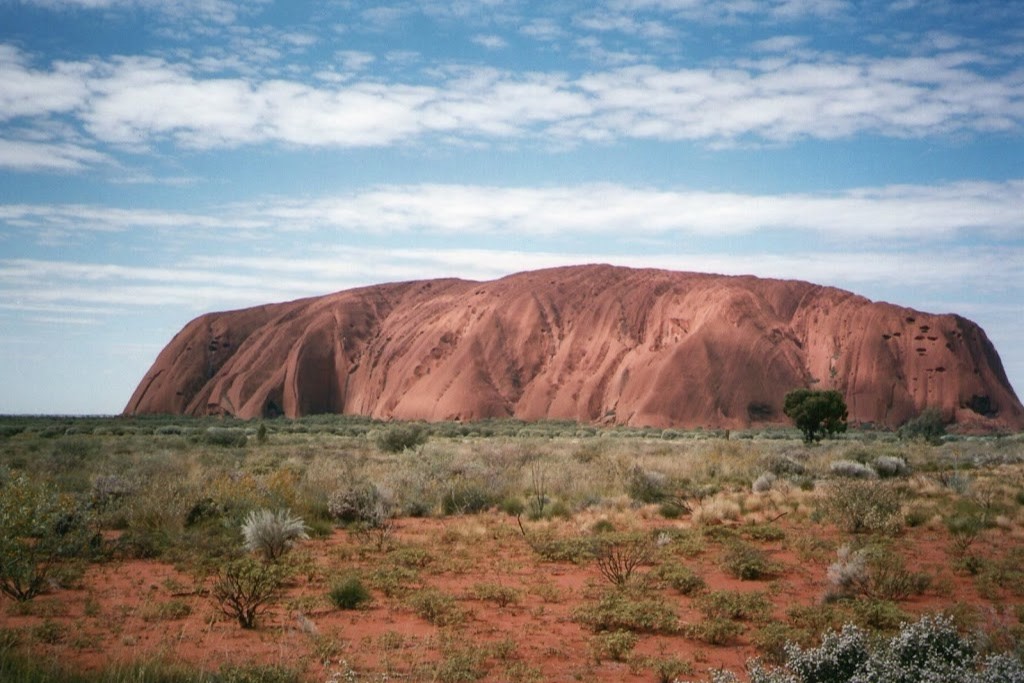 Uluru, also known as Ayers Rock, a massive sandstone monolith located in the arid landscape of the Northern Territory, Australia. The iconic rock formation stands prominently against a partly cloudy sky, surrounded by red soil and sparse desert vegetation. The scene captures the natural beauty and cultural significance of this famous landmark.