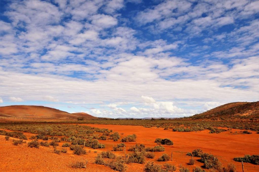 Wide-open outback landscape with rolling red hills, scattered shrubs, and a partly cloudy sky. The scene showcases the vastness and raw beauty of the Australian desert.