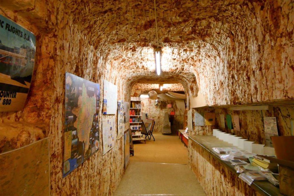 Underground bookshop in Coober Pedy, South Australia, with walls carved out of the rock, revealing a textured, reddish-brown surface. The narrow hallway is lined with posters and maps, leading to a small retail area with bookshelves and displays. The unique subterranean setting highlights the town's distinctive underground lifestyle and architecture.