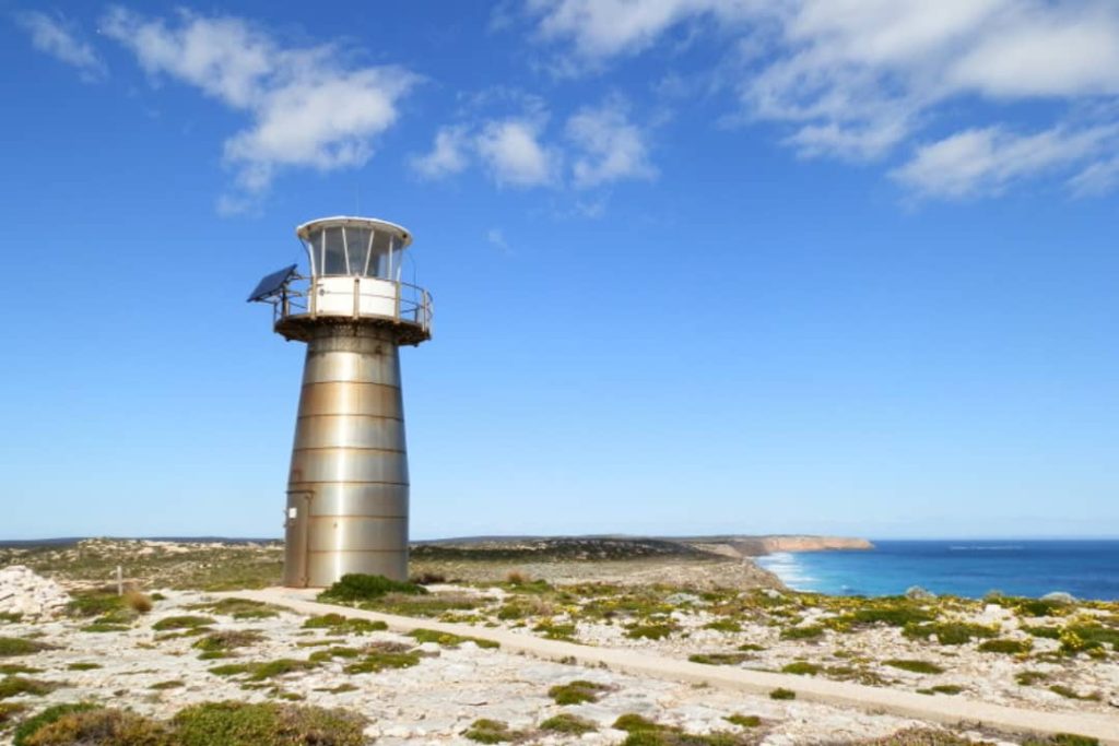 A sturdy metal lighthouse stands tall under a bright blue sky at West Cape in Innes National Park, South Australia. The scenic landscape features a rugged coastline with cliffs and a calm ocean in the distance, showcasing the beauty of the Yorke Peninsula.